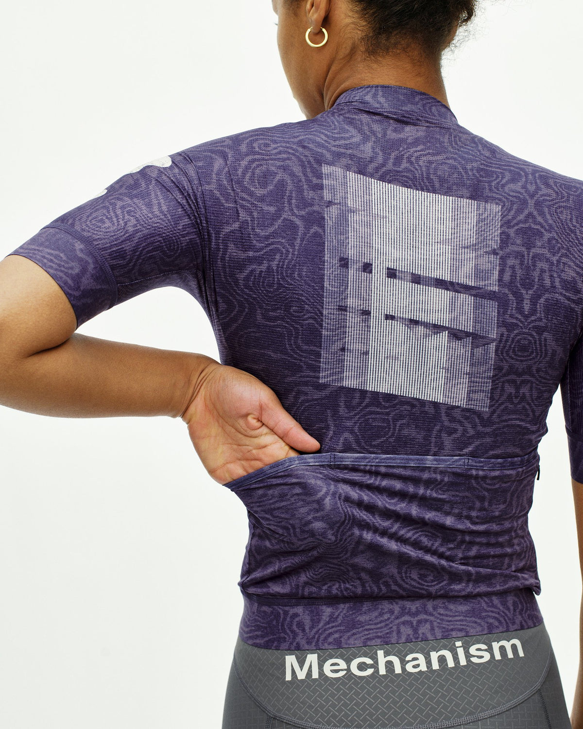 Maillot Corto Mujer Pas Normal Studios Mechanism Logo en AOP RTN Dark Purple, detalle de la espalda sobre ciclista #Color_AOP RTN Dark Purple