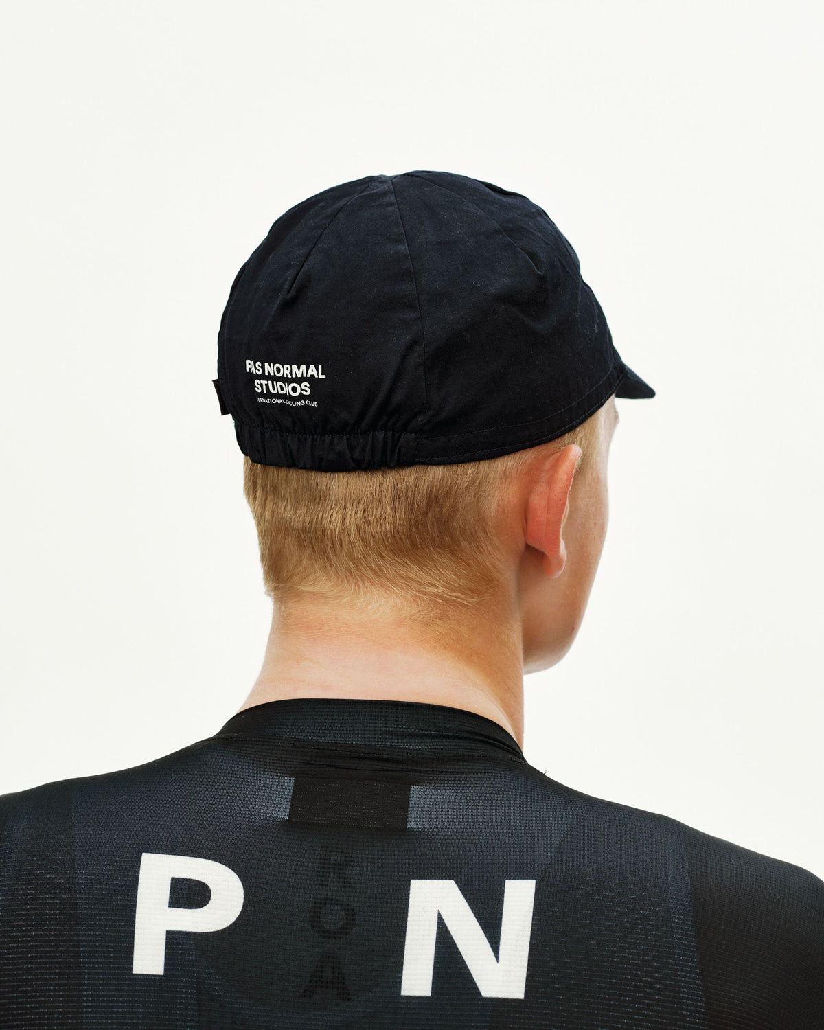 Black Logo Cap cycling cap, rear view on model with white logo print and elastic hem #Color_Black